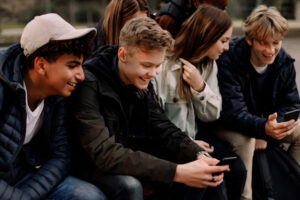 Smiling Teenage Girls And Boys Using Mobile Phones While Sitting At Park